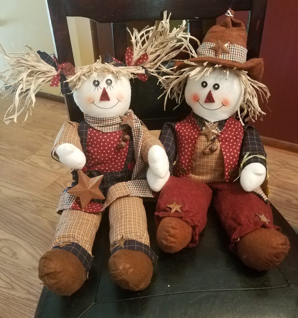 Two scarecrows sitting on a chair in the middle of room.