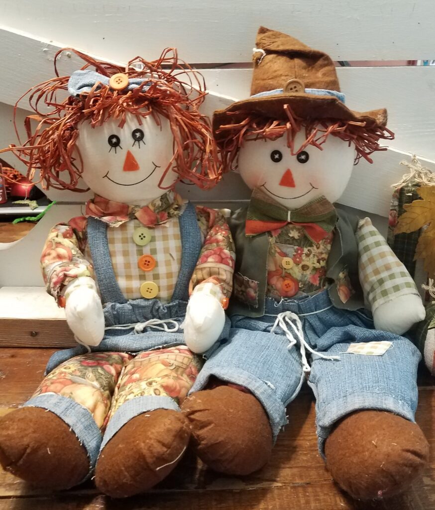 Two dolls are sitting on a table and one is wearing an overalls.