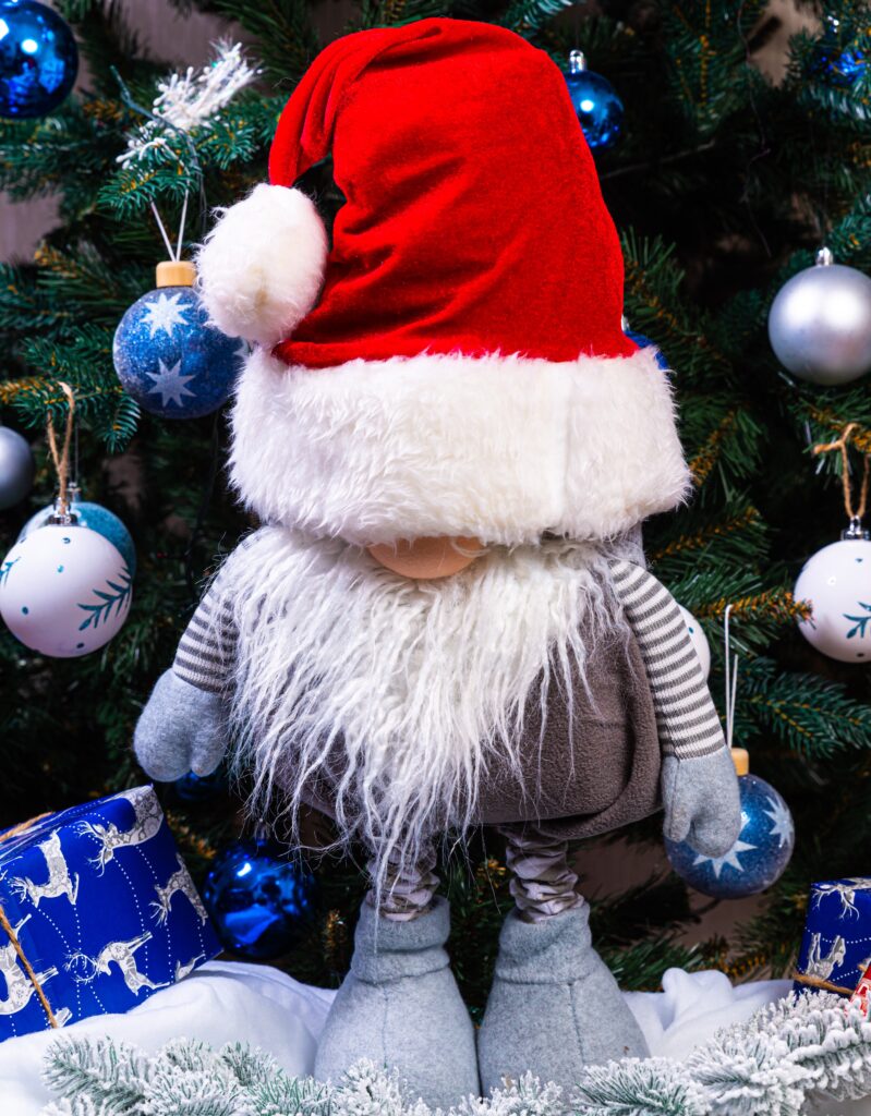 A gnome doll wearing a santa hat and standing in front of christmas tree.